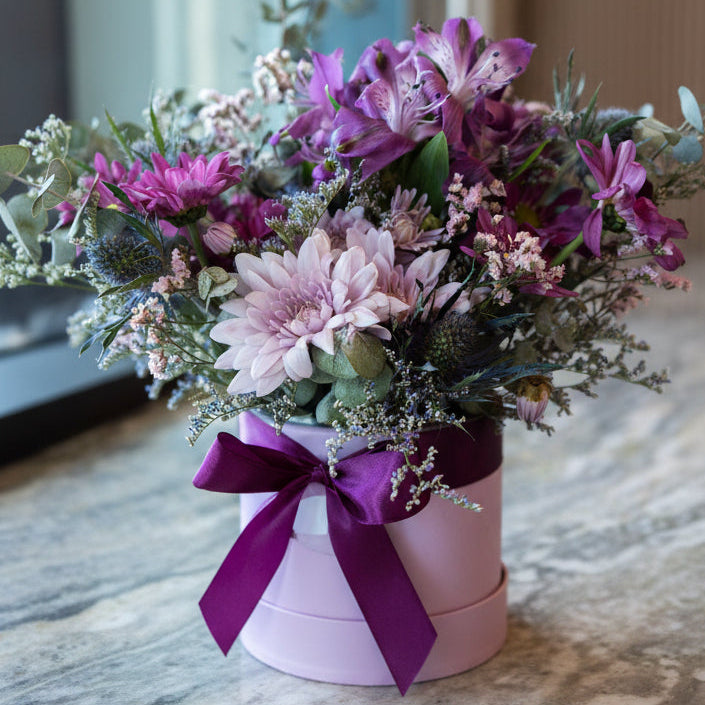 Bouquet of flowers in a pink container with a purple bow on a marble surface.